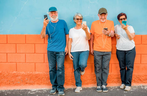 gruppo allegro di anziani appoggiati a un muro arancione e blu che indossano auricolari guardando i loro telefoni godendo della tecnologia e dei social - couple dependency standing men foto e immagini stock
