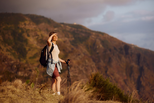 Smiling female hiker standing on a mountain and shielding her eyes while looking at distance.