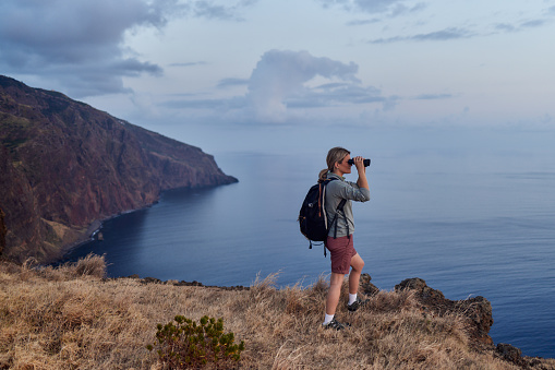 Young female hiker using binoculars while looking at distance from a cliff above the sea.