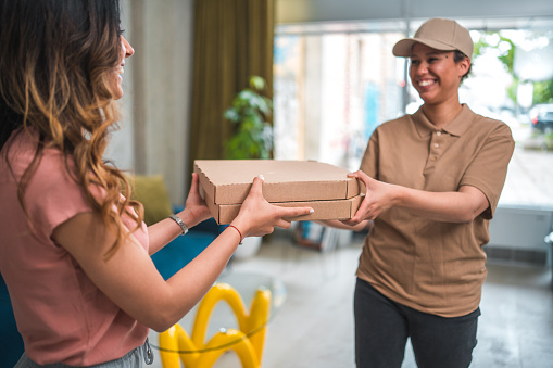 Dressed in professional attire, a mid adult Asian female employee receives a food delivery, expressing happiness and a warm smile. Engaging in conversation with her is a mixed race delivery woman.