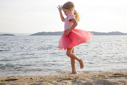 Cheerful little girl in pink dress dancing on the beach. Concept of freedom, happiness and positivity.