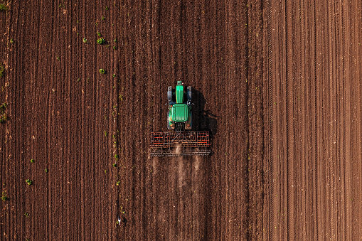 Green tractor vehicle with tiller attached performing field tillage before the sowing season, aerial shot seen from the drone pov top down