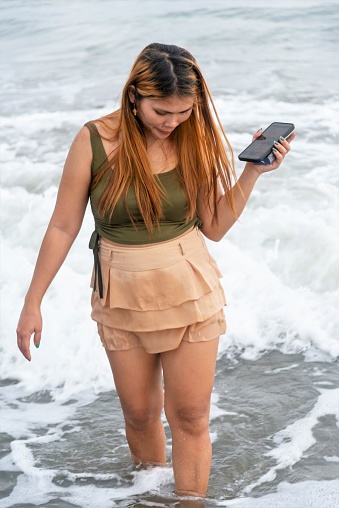 Holding her phone for selfies,on a beach near Manila at dusk,aimlessly strolling the warm shoreline waters,along smooth sands,having fun,dancing around,kicking the gentle waves as they roll in.