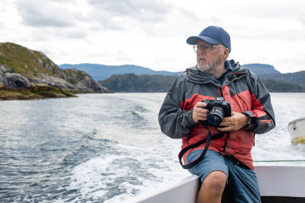 A senior man enjoying a tourist excursion while on holiday in Torridon, Scotland. He is sitting on a tourboat and holding a camera, ready to take pictures with it.