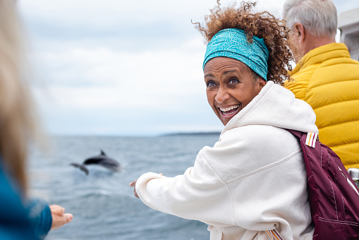 A group of friends enjoying a weekend away together in Torridon, Scotland, they are on a tourboat, enjoying an excursion out at sea. The main focus is one woman looking over her shoulder excitedly while pointing at a dolphin that is jumping out the water.