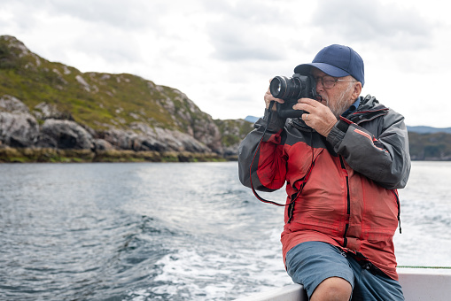 A senior man enjoying a tourist excursion while on holiday in Torridon, Scotland. He is on a tourboat and taking a photo of the view with his camera.