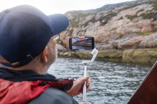 A man enjoying a tourist excursion while on holiday in Torridon, Scotland. He is on a tourboat and taking a video of the view using his phone and a gimbal to stabilise the shot.