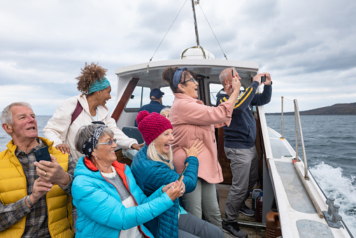 A group of friends enjoying a weekend away together in Torridon, Scotland. They are on a tourboat, enjoying an excursion out at sea while taking pictures with their phones of the view and looking excited.