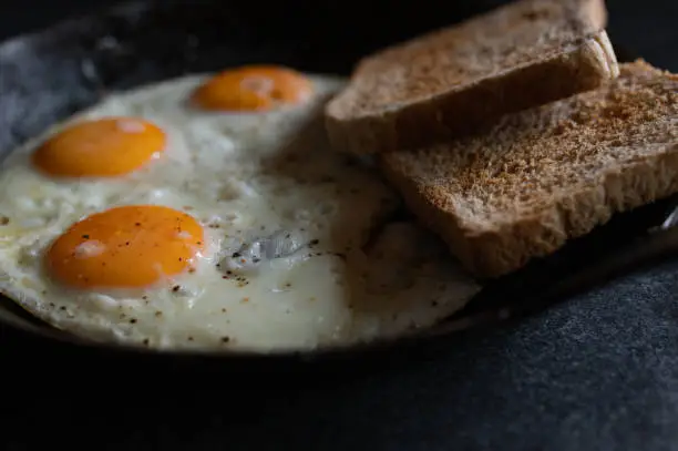 Fried eggs, sunny side up with pepper and salt seasoning whole wheat toast in a rustic cast iron pan on dark table background. Close up, front view