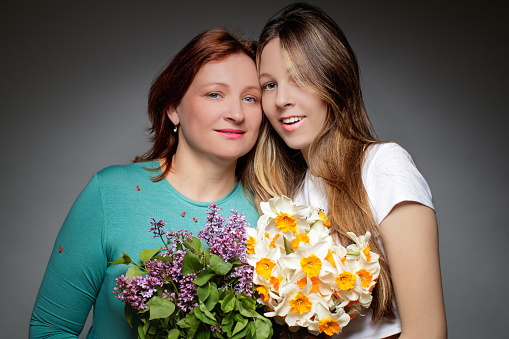 Middle aged woman with dyed red hair standing close to her naturally blond teenage daughter, they are smiling at camera and holding flower bunches, Mother's Day celebration, studio shot
