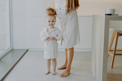 Cute girl in white robe poses with mom, hair combed, ponytail, bare feet. Haircare and children concept.