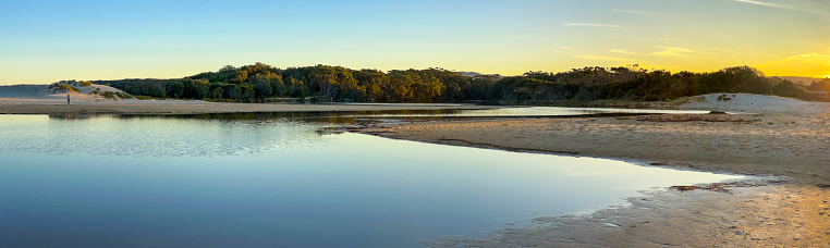 Horizontal panoramic view of the coastal forest, sand dunes and calm blue waters of beautiful Lake Tabourie lit up by the late afternoon sunset in Meroo National Park near Ulladulla, south coast NSW.
