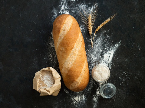 British White Bloomer or European sourdough Baton loaf bread on black background. Fresh loaf bread and glass jar with sourdough starter, floer in paper bag and ears. Top view. Copy space