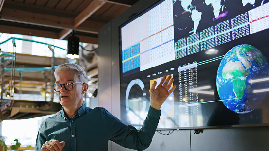 Stock image of a mature man conducting a seminar / lecture with the aid of a large screen. The screen is displaying graphs & data associated with images of the earth.