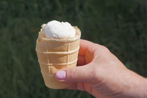 one white ice cream in a brown waffle cup is held by a girls hand on the street on a green background