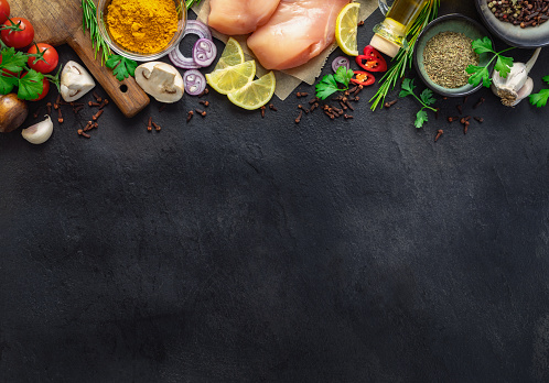 Food cooking ingredients background with fresh vegetables, chicken breast, herbs, spices and olive oil on dark stone table top view. Healthy diet eating