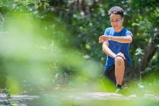 Asian man in huge blue t-shirt looking at smart watch, checking distance tracking while stretching in wood park with smile of happiness, copy space at left.