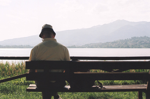 A Senior Man Sitting and Reading on the Pusiano Lake Park Bench during a Sunny Summer Day. Lecco Province, Italy. Film Photography