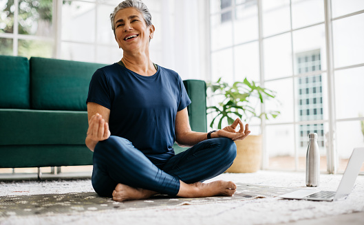 Smiling senior woman meditating at home, she is sitting in front of a laptop in lotus position, following a virtual yoga class. Mature woman practicing yoga for her mental and physical wellness.