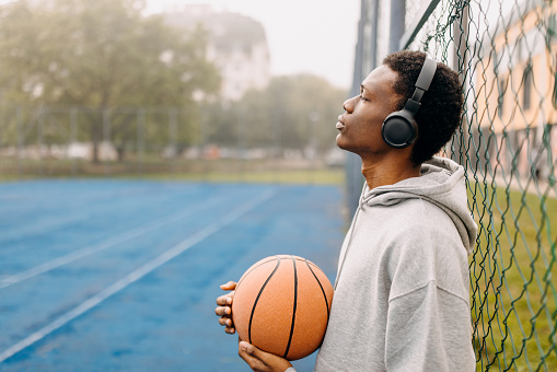 Young black basketball player with wireless headphones holding basketball in hands at sports court, leaning the fence