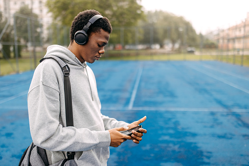 Young black athlete with wireless headphones holding gym bag at sports court, using his smart phone while walking