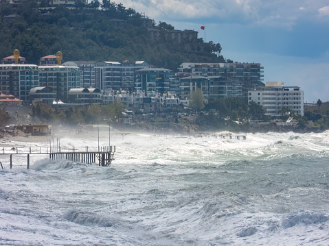 Alanya, Turkey - Mar 23, 2023. Rough waves crashing into the coast with various buildings situated nearby on the shore. Stormy Mediterranean sea.