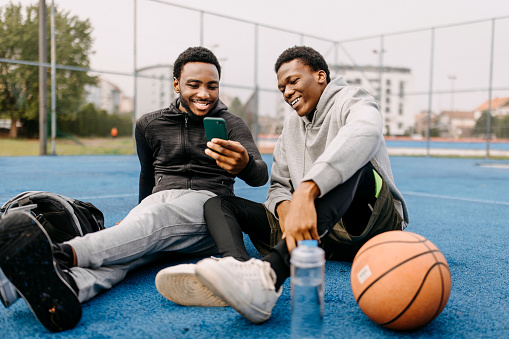 Two young smiling friends using phone while sitting on a ground at outdoor basketball court, surfing the internet.