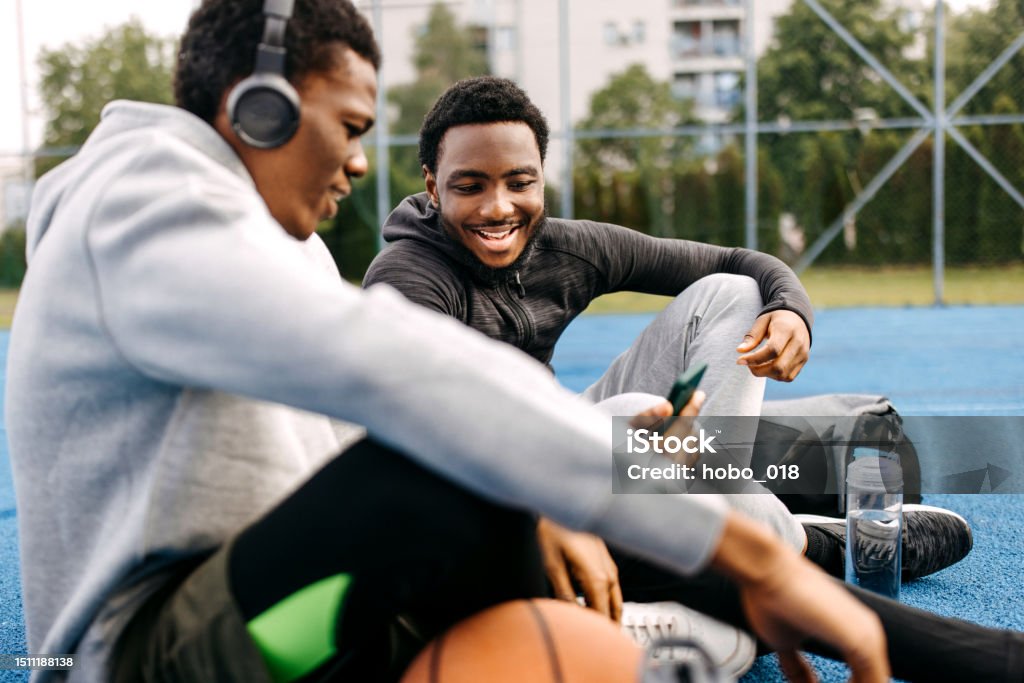 Two friends hanging together on basketball court, using smart phone Two young smiling friends using phone while sitting on a ground at outdoor basketball court, listening to the music Basketball - Ball Stock Photo