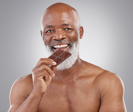 Black man, senior portrait and eating chocolate isolated on a  studio background for a treat. Happy, snack smile and an elderly African model biting into a sweet candy bar for happiness and sugar