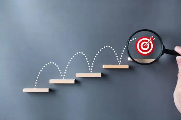 Photo of Unlock potential of business success stairs dart and dartboard targets magnifying glass with hand on gray background. Explore opportunities growth embrace steps to achieve ambitions and goal concept.