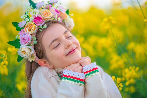 Dreaming Beautiful young girl with flower chaplet, ethnic folklore dress with traditional Bulgarian embroidery during sunset on a rapseed agricultural field
