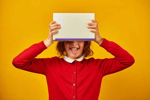 Portrait of happy teenage schoolgirl cover her face by book on yellow studio background, educatin concept, having fun , Back to School, copy space.