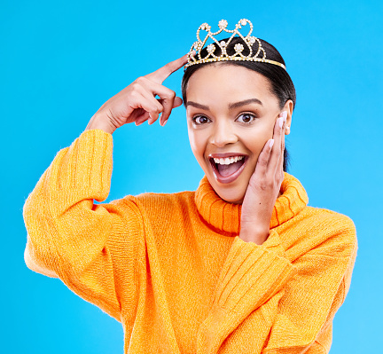 Pointing, crown and portrait of woman in studio for celebration, princess and party. Smile, beauty and fashion with female and tiara on blue background excited for achievement, winner and prom event