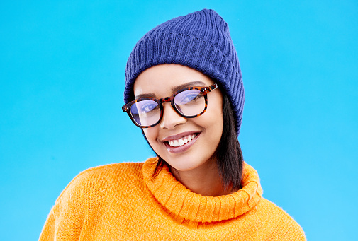 Portrait of woman in winter fashion with smile, beanie and glasses isolated on blue background. Style, happiness and gen z girl in studio backdrop with happy face and warm clothing for cold weather.