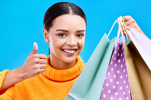 Happy woman, portrait smile and shopping bags with thumbs up for purchase, sale or discount against blue studio background. Face of female shopper with gift bag showing thumb emoji, yes sign or like
