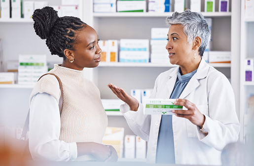 Female African American pharmacist holding bottle of skin cream talking to senior woman about skin care routine.