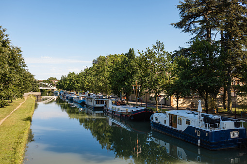 Wide shot of a canal in Toulouse in the South of France. There are moored riverboats next to the trees.