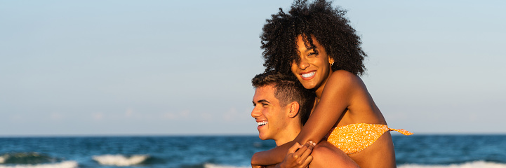 Happy young interracial couple in piggybak on beach – man giving piggybak his woman and laughing at tropical beach – smiling boyfriend in love carrying on back her girlfriend and having fun  - banner size for header design