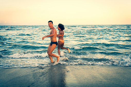 multiracial young couple running in shoreline – diverse two friends run on beach – multicultural people tourist in vacation in summer