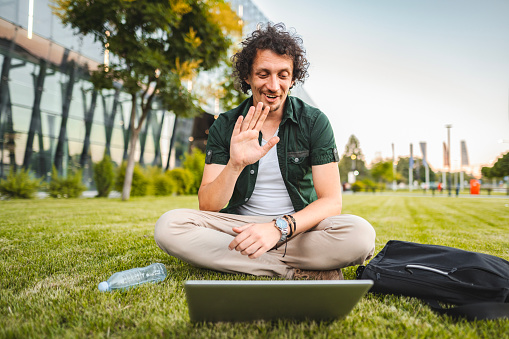 Men talking on a video call on his laptop while sitting on the grass in public park