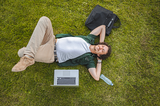 Men lying on the grass in public park while taking a break from working on his laptop
