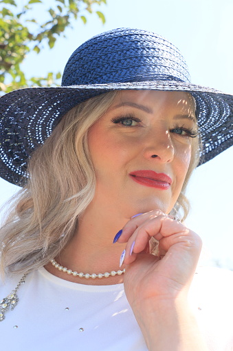 A closeup of a blond haired Irish woman. She is wearing medium length blond hair, makeup, a blue straw hat, necklace, false nails and a white top.