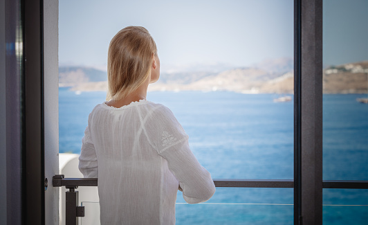 Rear view on a dreamy woman enjoying beautiful view of mountains behind the sea from her hotel room. Spending time on open air terrace. Travel and vacation.