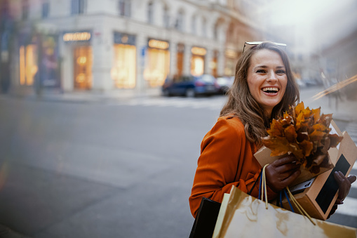Hello november. smiling stylish woman in brown trench coat with parcels, shopping bags and autumn yellow leaves in the city.