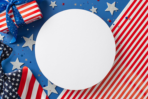 Honoring USA's Independence Day. Top view captures essence of celebration: glittering stars, confetti, tie, giftbox wrapped in thematic paper, American flag backdrop with vacant circle for text or ad