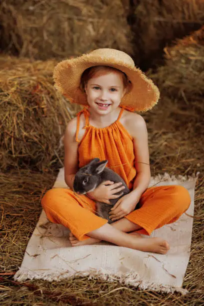 Photo of Little cute girl in an orange dress and a straw hat sits on straw bales with a brown rabbit