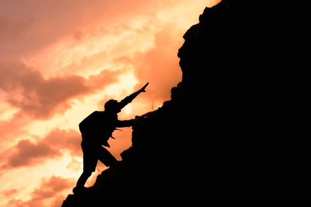 Caucasian female athletic blond rock climber on the sheer face of a granite cliff reaching up with her hands to gain more heights.  Subject is wearing all the appropriate climbing gear such as belts and carabiners to manage the safety rope.  Climber and cliff is positioned perfectly at the right third of the image revealing a pine tree forest in the distance creating ample copy space for titles and other design elements.