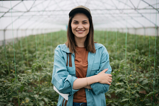 Portrait of a happy young greenhouse farm owner. Female farmer with arms crossed looking at camera and smiling in tomato farm in greenhouse.