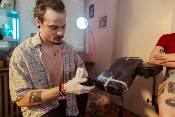 Portrait of a young Cuban man sitting in his tattoo studio and focusing on cleaning a machine. His client is waiting on the tattoo chair.