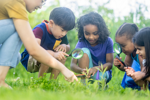 A female teacher of Asian decent, takes her class outside as they discover the world around them through magnifying glasses.  They are each dressed casually and are bright-eyed as they look for insects and bugs during their science class exploration.